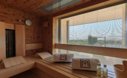 Interior of a modern sauna featuring wooden benches, towels with a logo, a clock, and a view through large windows with blinds. The space is designed for relaxation, with essential oils placed on a bench for aromatherapy.