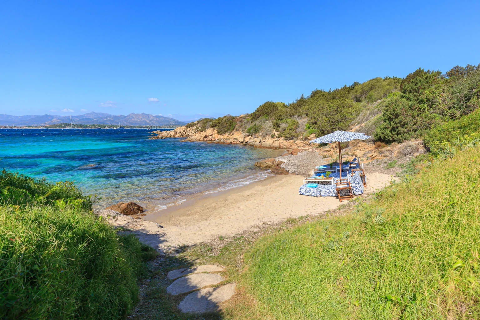 Scenic beach view featuring turquoise waters, sandy shore, and lounging area with umbrellas and chairs, surrounded by lush greenery and rocky coastline under a clear blue sky. Ideal for relaxation and outdoor activities. | Only on SopranoVillas