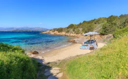 Scenic beach view featuring turquoise waters, sandy shore, and lounging area with umbrellas and chairs, surrounded by lush greenery and rocky coastline under a clear blue sky. Ideal for relaxation and outdoor activities.