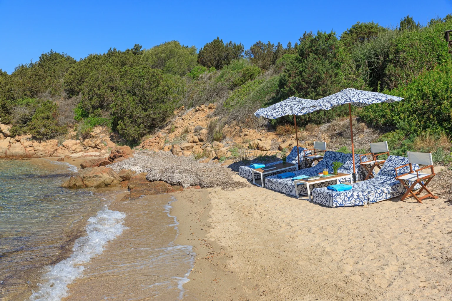 Relaxing beach scene featuring lounge chairs with patterned cushions under umbrellas, set against a backdrop of sandy shore and rocky coastline, surrounded by lush greenery and clear blue skies. Perfect destination for a tranquil getaway or sunbathing experience. | Only on SopranoVillas
