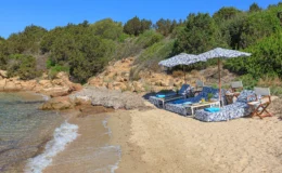 Relaxing beach scene featuring lounge chairs with patterned cushions under umbrellas, set against a backdrop of sandy shore and rocky coastline, surrounded by lush greenery and clear blue skies. Perfect destination for a tranquil getaway or sunbathing experience.