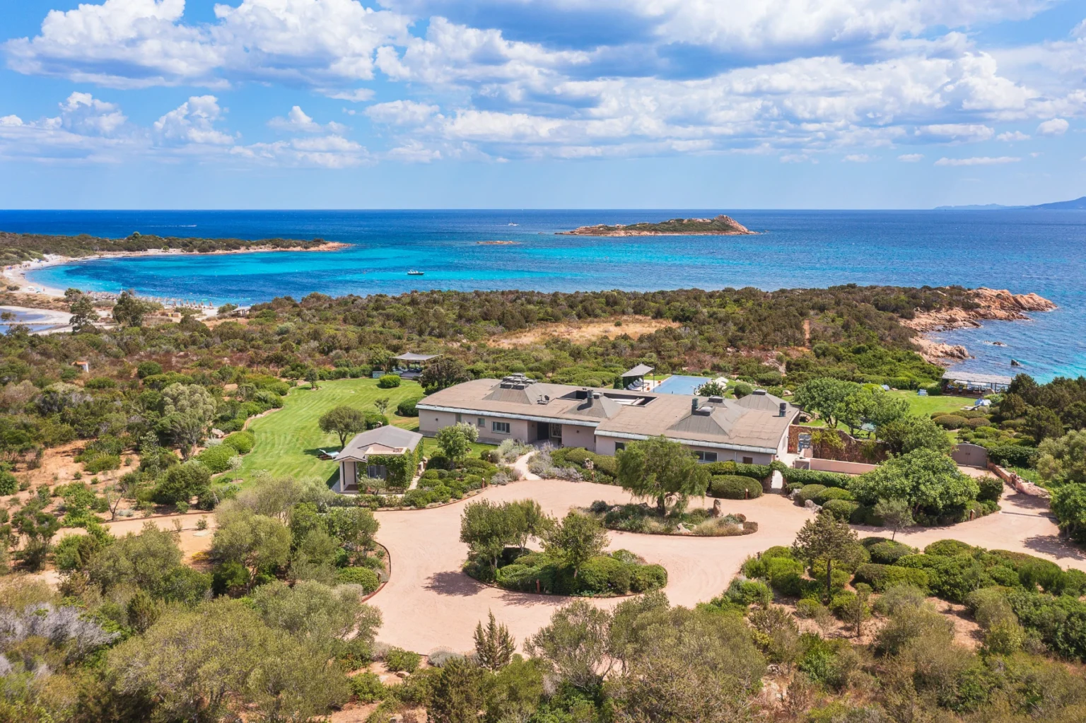 Aerial view of a luxurious coastal property surrounded by lush greenery, featuring a spacious lawn, landscaped gardens, and a private beach area with turquoise waters and rocky islands in the background. | Only on SopranoVillas