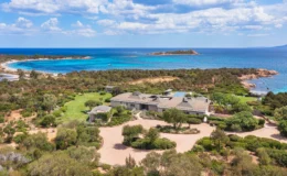 Aerial view of a luxurious coastal property surrounded by lush greenery, featuring a spacious lawn, landscaped gardens, and a private beach area with turquoise waters and rocky islands in the background.