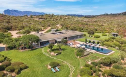 Aerial view of a luxurious modern villa surrounded by lush greenery and mountains, featuring a spacious lawn, a swimming pool with lounge chairs, and beautifully landscaped gardens. The image showcases the property's outdoor living spaces and natural beauty.