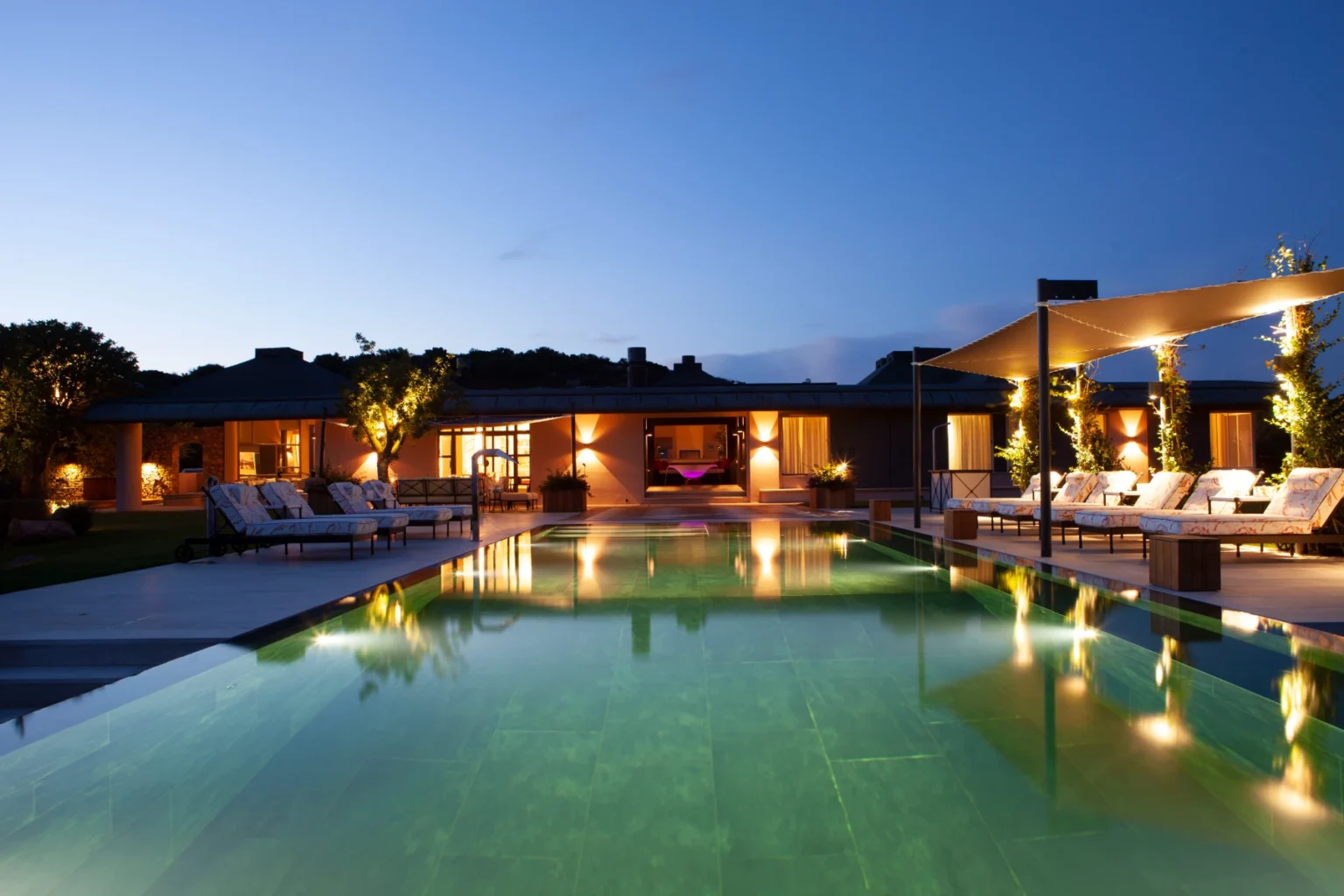 Luxurious modern villa with a serene swimming pool surrounded by lounge chairs and illuminated by soft lighting at dusk, showcasing a tranquil outdoor space perfect for relaxation and entertainment. | Only on SopranoVillas