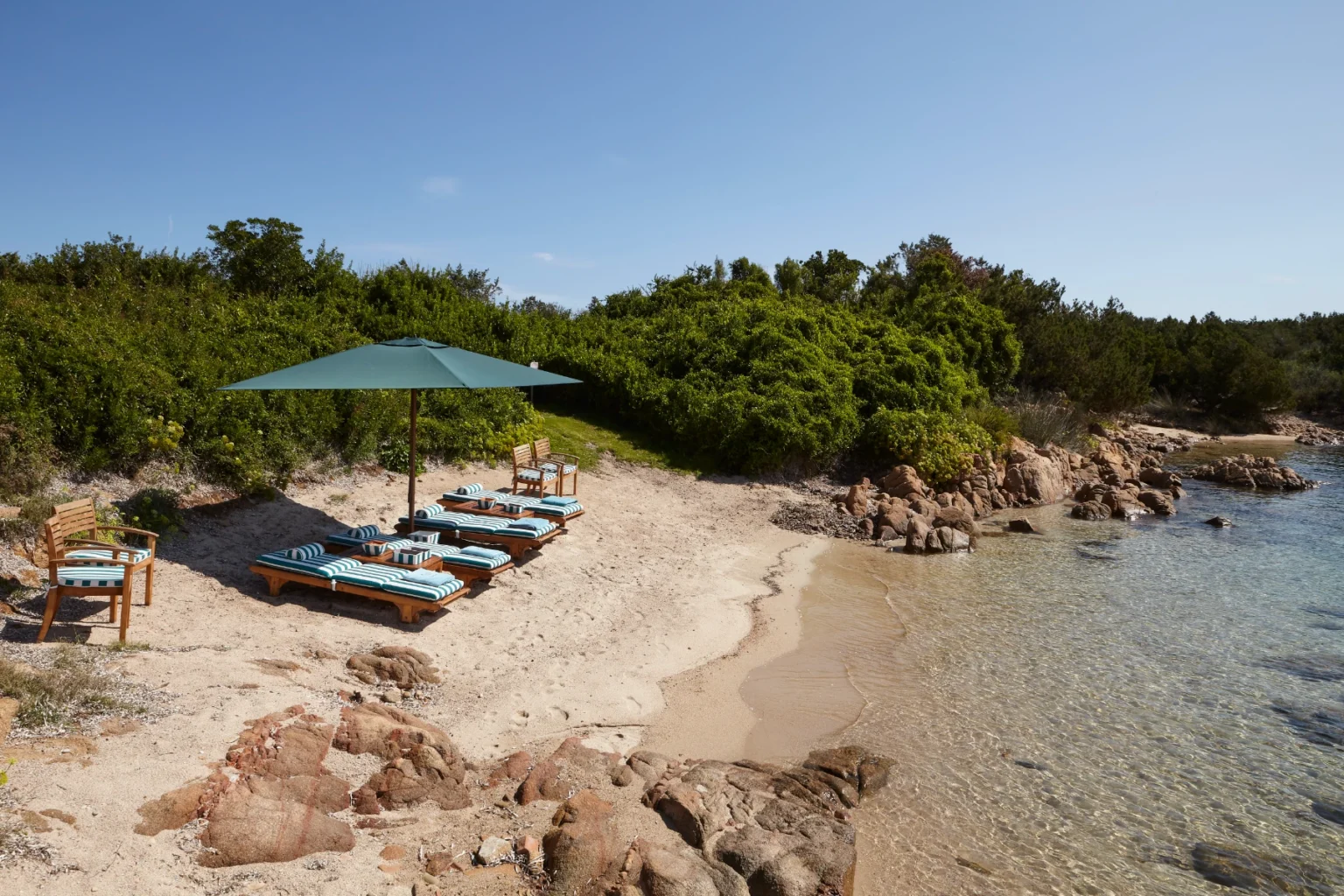 Beach scene featuring lounge chairs with blue and white striped cushions under a green umbrella, surrounded by sandy shore and lush greenery, with clear water lapping at the shore. Ideal for relaxation and sunbathing. | Only on SopranoVillas
