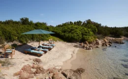 Beach scene featuring lounge chairs with blue and white striped cushions under a green umbrella, surrounded by sandy shore and lush greenery, with clear water lapping at the shore. Ideal for relaxation and sunbathing.
