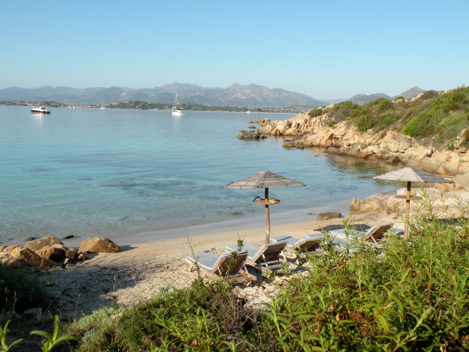 Serene beach scene with lounge chairs and umbrellas, clear turquoise water, rocky coastline, and distant mountains under a blue sky, ideal for relaxation and scenic views. | Only on SopranoVillas