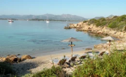 Serene beach scene with lounge chairs and umbrellas, clear turquoise water, rocky coastline, and distant mountains under a blue sky, ideal for relaxation and scenic views.