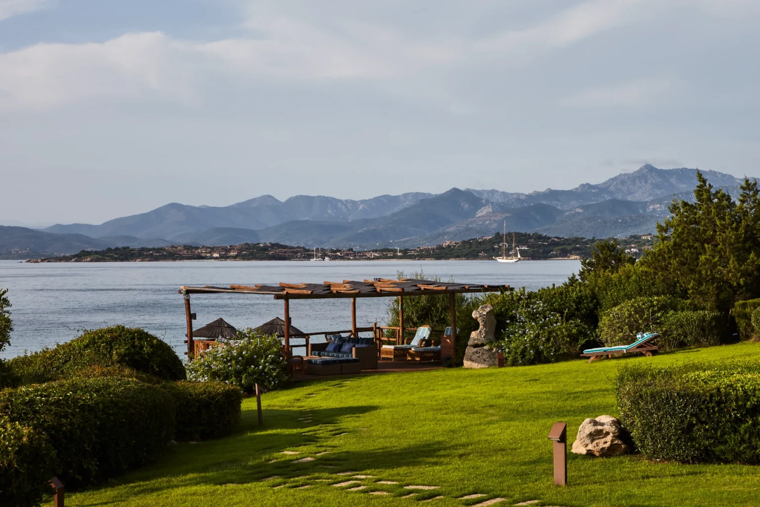 Scenic view of a coastal landscape featuring lush green grass, wooden seating area, and distant mountains, with a calm sea and a sailboat in the background. Ideal for relaxation and outdoor enjoyment. | Only on SopranoVillas