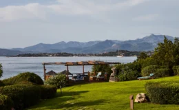 Scenic view of a coastal landscape featuring lush green grass, wooden seating area, and distant mountains, with a calm sea and a sailboat in the background. Ideal for relaxation and outdoor enjoyment.