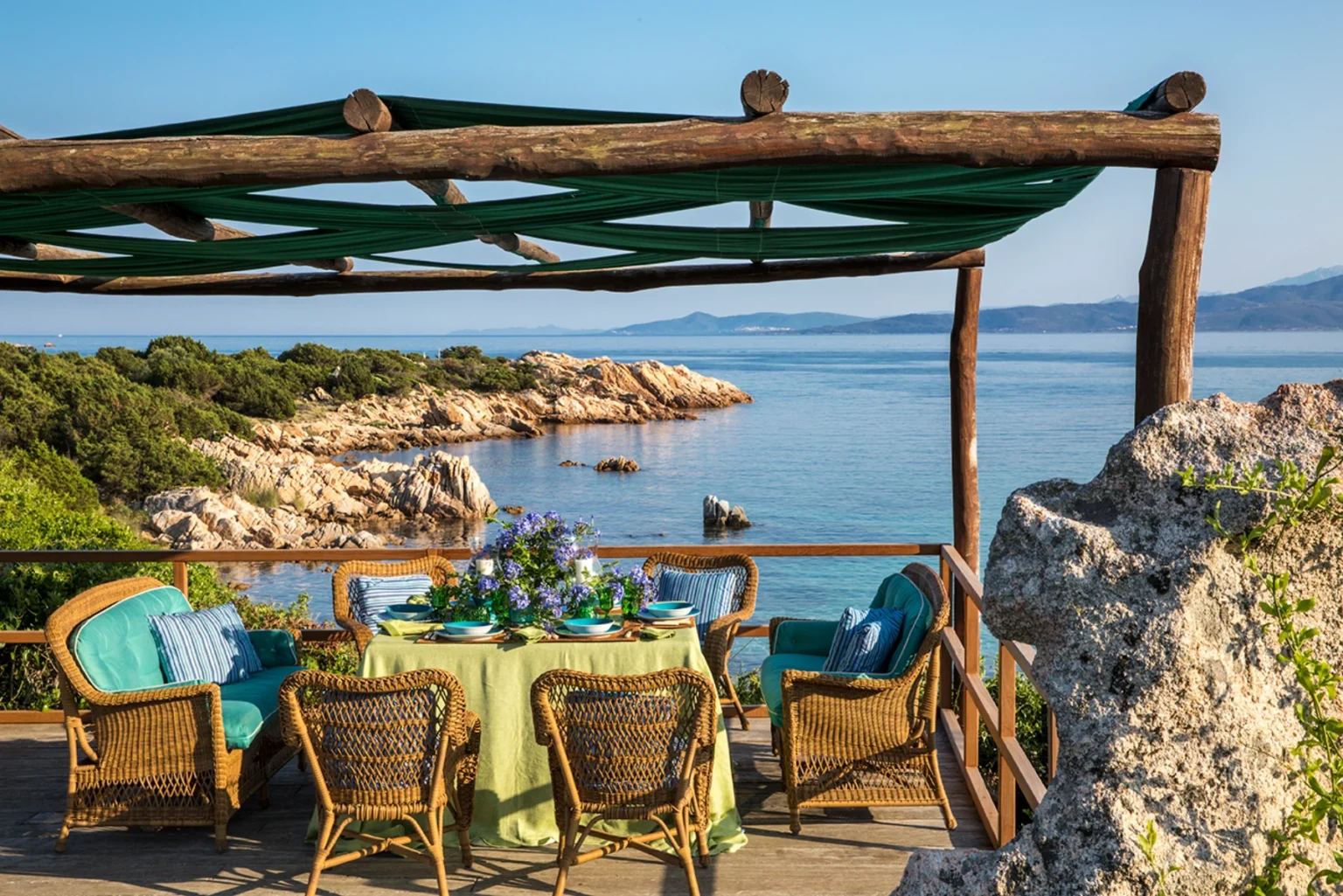 Scenic outdoor dining area overlooking a tranquil sea, featuring a wooden pergola with green fabric, a table set with floral arrangements, and comfortable woven chairs, surrounded by lush greenery and rocky coastline. | Only on SopranoVillas
