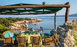 Scenic outdoor dining area overlooking a tranquil sea, featuring a wooden pergola with green fabric, a table set with floral arrangements, and comfortable woven chairs, surrounded by lush greenery and rocky coastline.