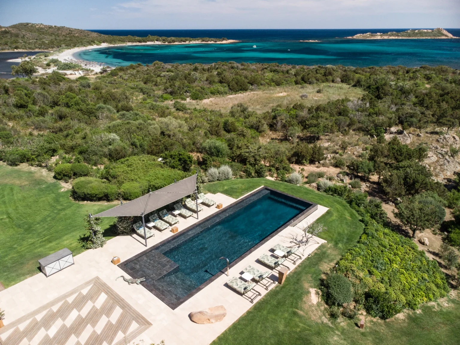 Aerial view of a luxurious outdoor pool surrounded by lush greenery, featuring comfortable lounge chairs and a shaded area. The scene overlooks a stunning coastline with turquoise waters and sandy beaches, ideal for relaxation and leisure. | Only on SopranoVillas