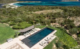Aerial view of a luxurious outdoor pool surrounded by lush greenery, featuring comfortable lounge chairs and a shaded area. The scene overlooks a stunning coastline with turquoise waters and sandy beaches, ideal for relaxation and leisure.