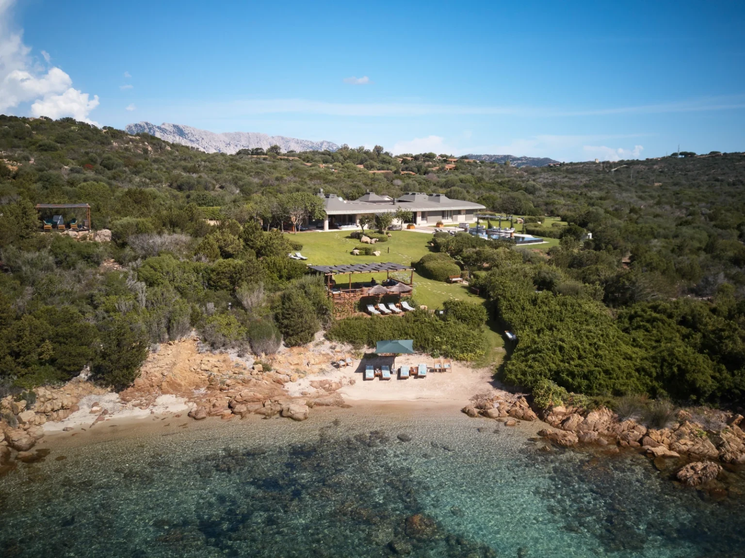 Aerial view of a luxurious beachfront villa surrounded by lush greenery and rocky shoreline, featuring landscaped gardens, outdoor seating areas, and a clear turquoise sea. Ideal for vacation rentals or private getaways in a serene natural setting. | Only on SopranoVillas