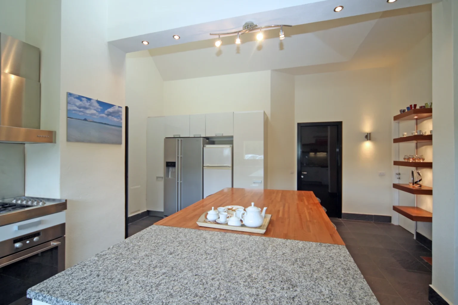 Modern kitchen design featuring a large granite island, sleek stainless steel appliances, and minimalist cabinetry. The space is illuminated by recessed lighting and includes a decorative shelf with glassware, creating a stylish and functional cooking area. | Only on SopranoVillas