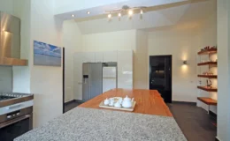 Modern kitchen design featuring a large granite island, sleek stainless steel appliances, and minimalist cabinetry. The space is illuminated by recessed lighting and includes a decorative shelf with glassware, creating a stylish and functional cooking area.