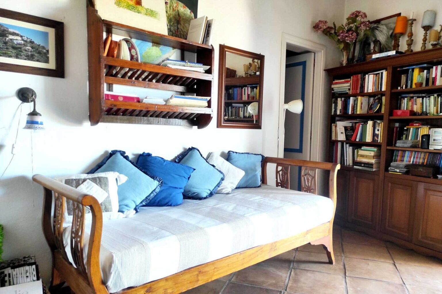 Cozy living room featuring a wooden daybed adorned with blue cushions, surrounded by bookshelves filled with books and decorative items, creating a warm and inviting atmosphere. | Only on SopranoVillas