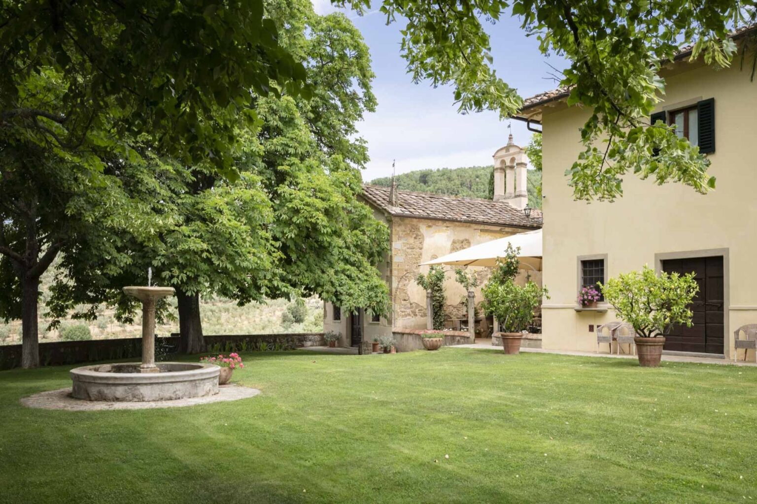 Charming outdoor view of a well-maintained garden featuring a stone fountain, lush green grass, and a rustic villa surrounded by trees, ideal for relaxation and outdoor gatherings. | Only on SopranoVillas