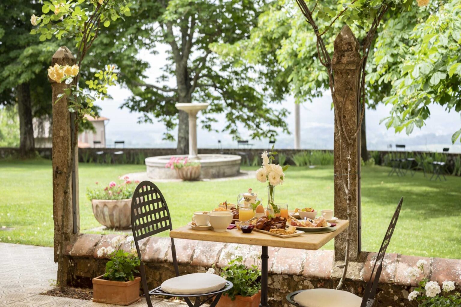 Outdoor dining setup in a lush garden featuring a wooden table adorned with an assortment of food, surrounded by greenery and a stone fountain in the background. Ideal for a relaxing meal in nature. | Only on SopranoVillas