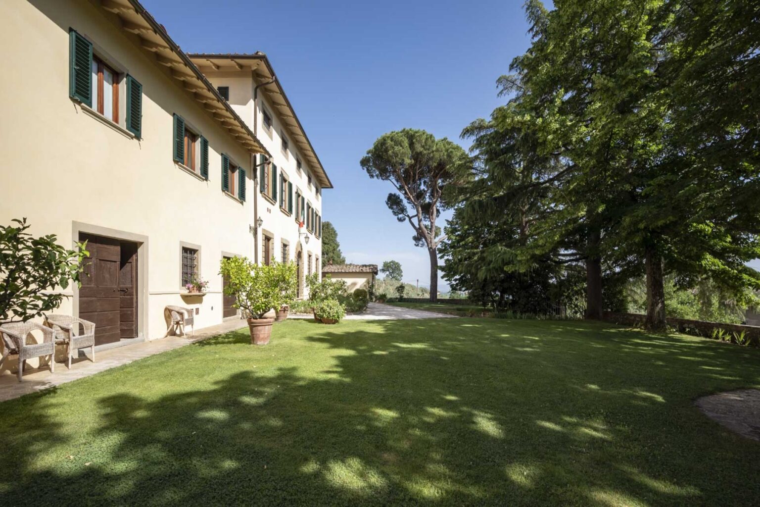 Tuscan villa with a manicured lawn, surrounded by trees and a clear blue sky, showcasing the tranquil outdoor space ideal for relaxation and gatherings. | Only on SopranoVillas