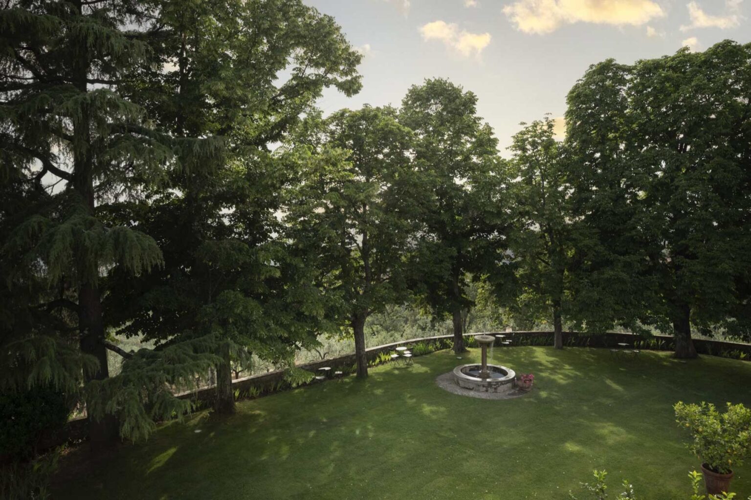 Lush green garden with tall trees surrounding a stone fountain, featuring manicured grass and a scenic view, ideal for relaxation and outdoor gatherings. | Only on SopranoVillas