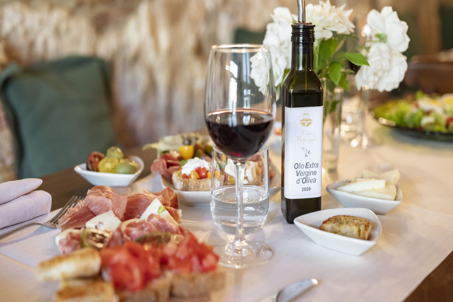 A beautifully arranged dining table featuring a selection of gourmet appetizers, including cured meats and fresh vegetables, complemented by a glass of red wine and a bottle of Italian olive oil, with a floral centerpiece adding elegance to the setting. | Only on SopranoVillas