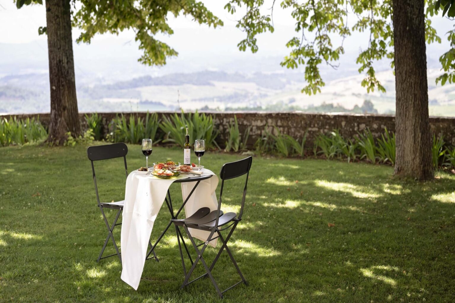 Cozy outdoor dining setup featuring a small table with wine glasses and a plate of food, surrounded by lush greenery and overlooking a scenic landscape. Ideal for romantic picnics or intimate gatherings in nature. | Only on SopranoVillas