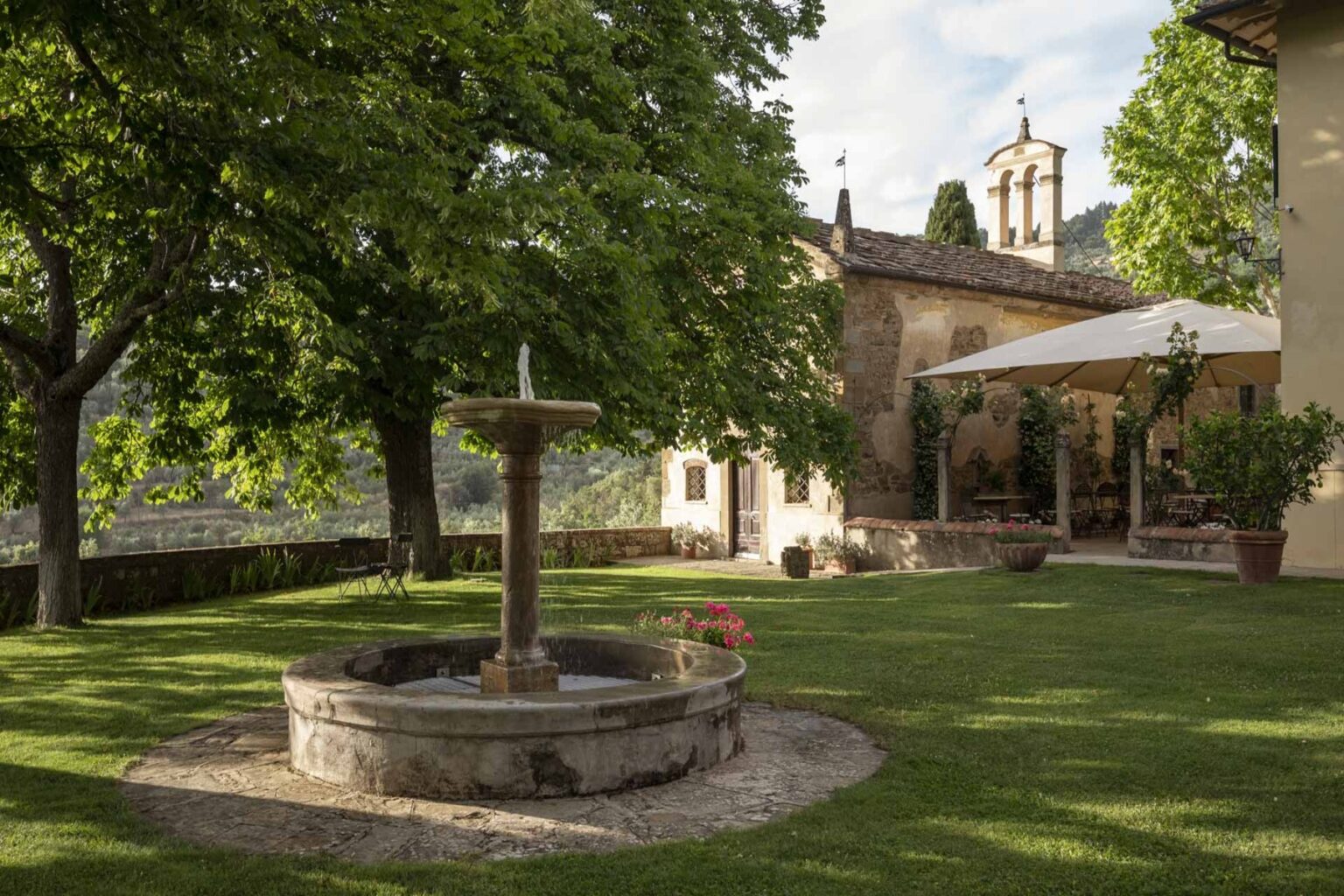 Scenic garden view featuring a stone fountain surrounded by lush greenery and trees, with a rustic building in the background and a shaded outdoor seating area, perfect for relaxation and gatherings. | Only on SopranoVillas