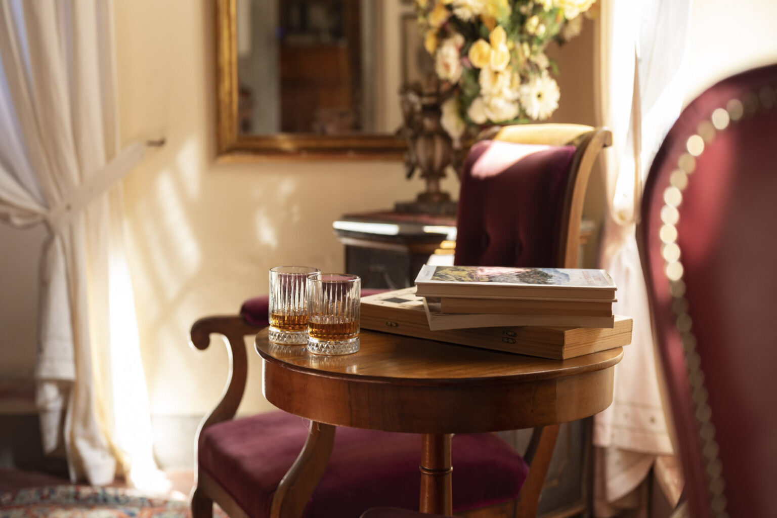 Elegant living room scene featuring a wooden side table with two glasses and a stack of books, complemented by a plush burgundy chair and a mirror reflecting soft natural light. | Only on SopranoVillas