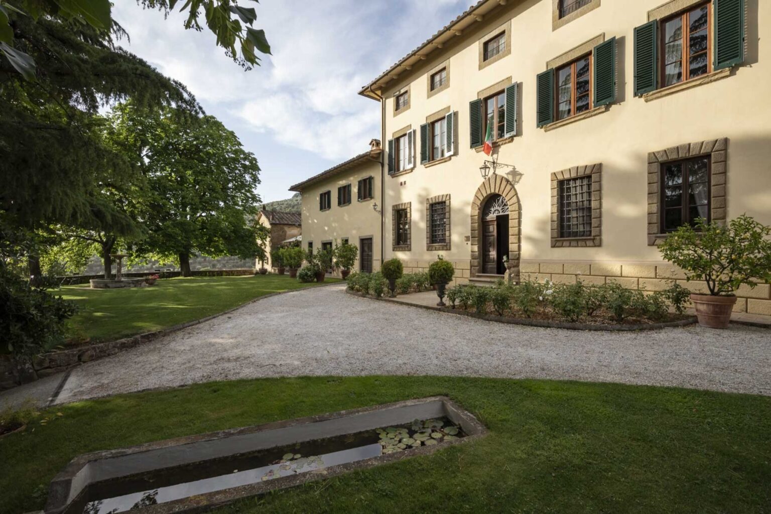 Historic villa exterior with lush green trees and manicured gardens, featuring a gravel pathway leading to a charming entrance with decorative windows and a stone façade. The scene captures a serene outdoor setting, ideal for relaxation and scenic views. | Only on SopranoVillas
