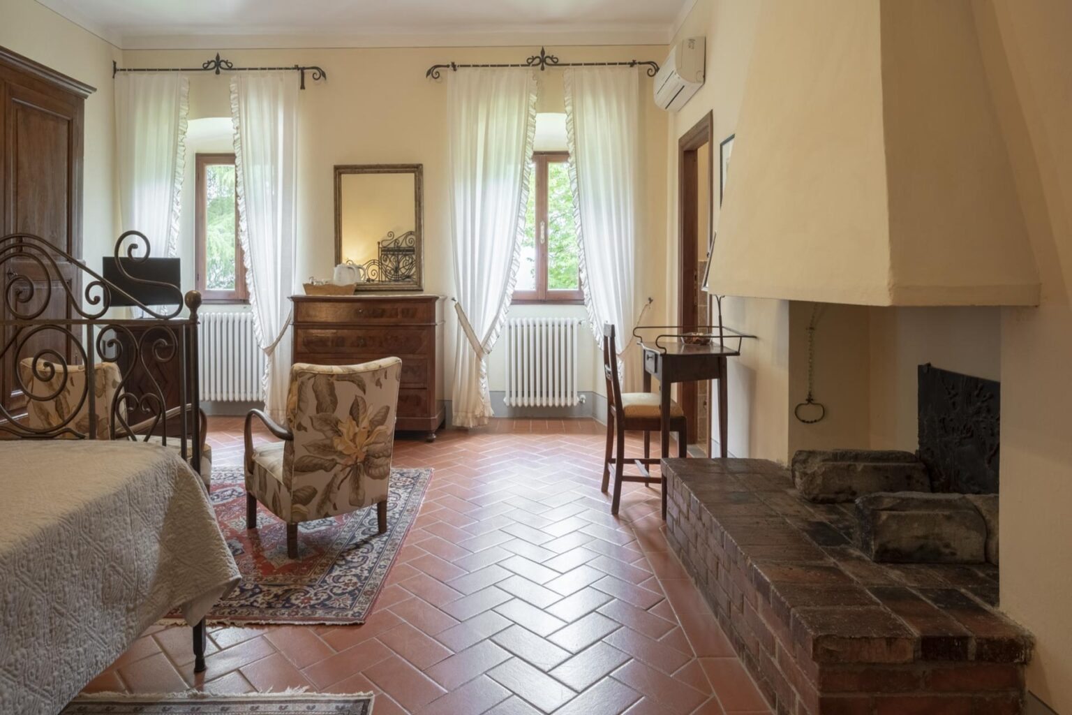 Cozy and elegant bedroom featuring a wrought iron bed, floral armchairs, wooden dresser, and a rustic fireplace. Natural light filters through sheer curtains, enhancing the warm ambiance of the terracotta tiled floor and charming decor. Ideal for a relaxing getaway. | Only on SopranoVillas