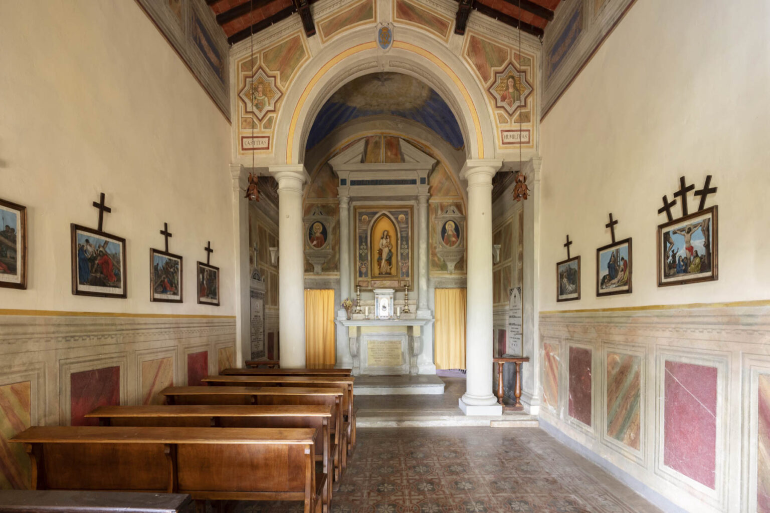 Interior view of a historic chapel featuring wooden pews, decorative wall paintings, and a central altar with an ornate backdrop. The chapel is adorned with framed religious artwork and crosses, showcasing its architectural and artistic significance. Soft lighting enhances the serene atmosphere of the space. | Only on SopranoVillas