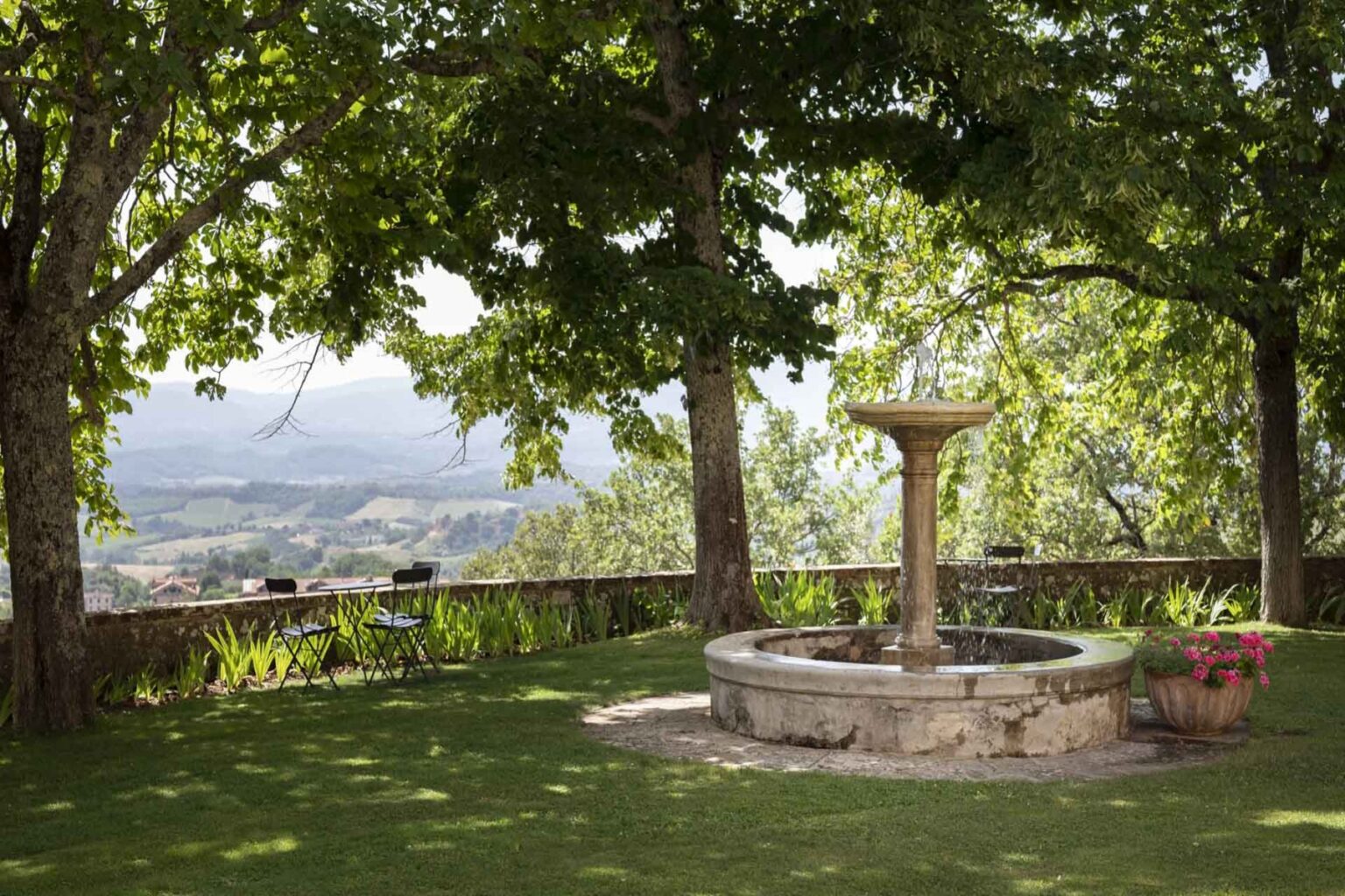 Scenic garden view featuring a stone fountain surrounded by lush greenery, trees, and a seating area with black chairs. The background showcases rolling hills and a tranquil landscape, ideal for relaxation and outdoor gatherings. | Only on SopranoVillas