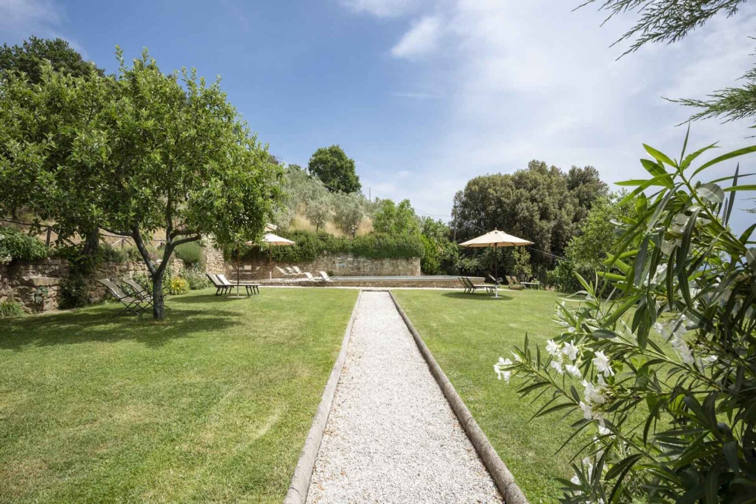 Lush garden pathway leading to a serene outdoor seating area with sun loungers and umbrellas, surrounded by trees and greenery under a clear blue sky. | Only on SopranoVillas