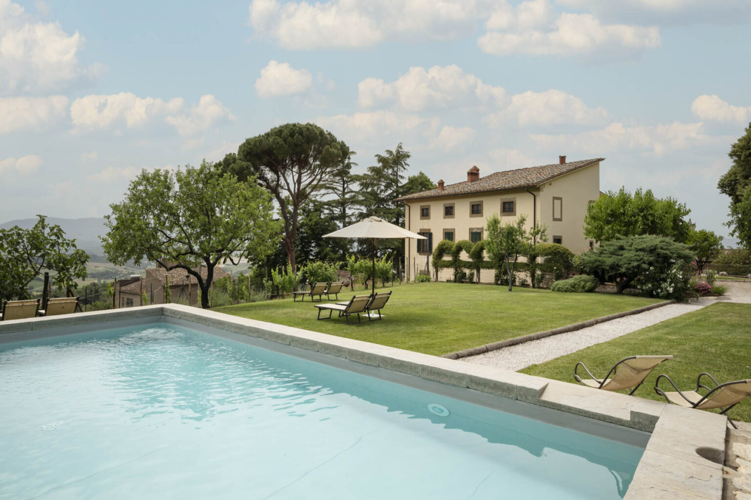 Scenic view of a luxurious villa with a swimming pool, surrounded by lush greenery and a well-maintained garden under a partly cloudy sky. | Only on SopranoVillas