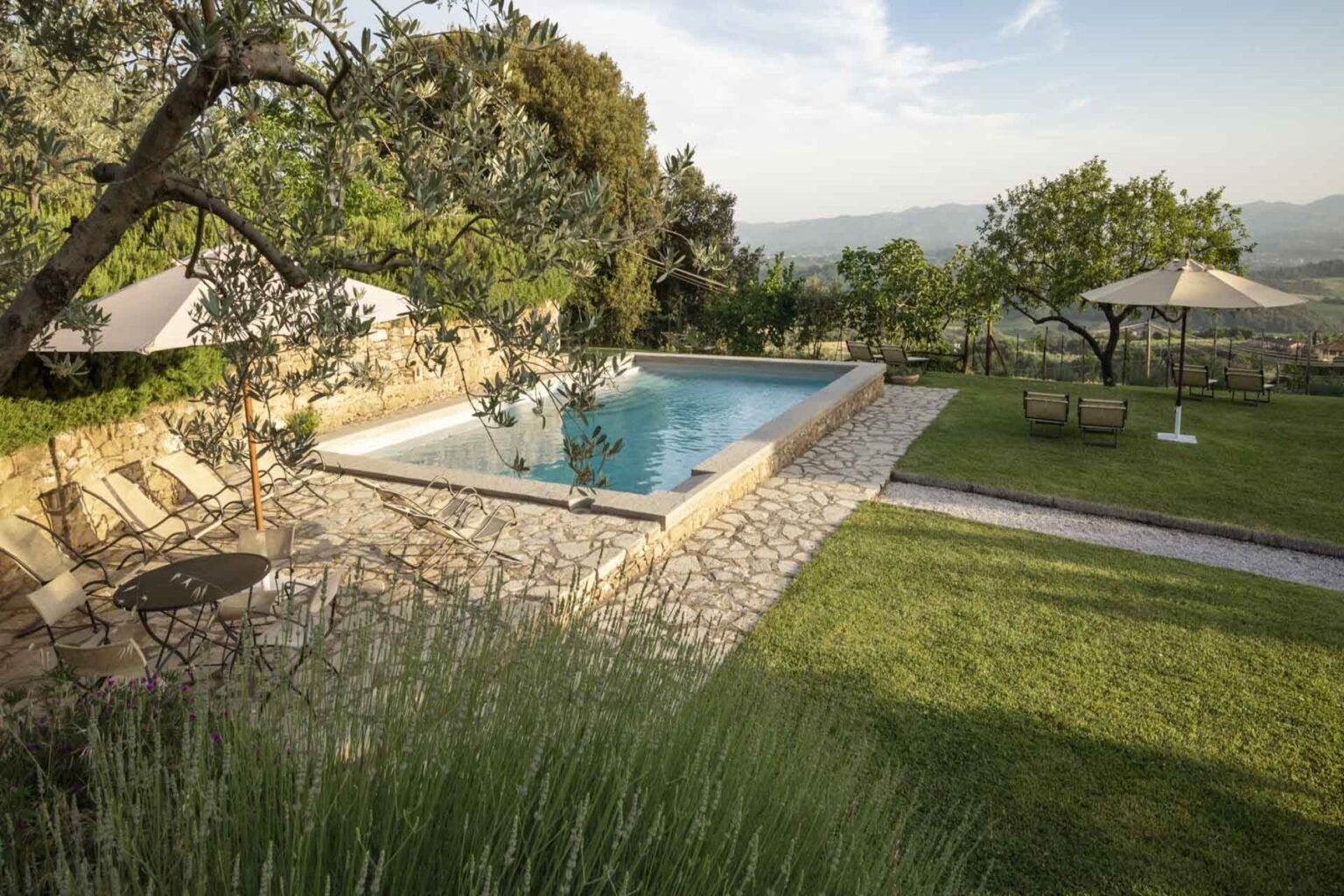 Scenic view of a tranquil outdoor swimming pool surrounded by lush greenery, stone pathways, and shaded seating areas, ideal for relaxation and leisure in a serene natural setting. | Only on SopranoVillas