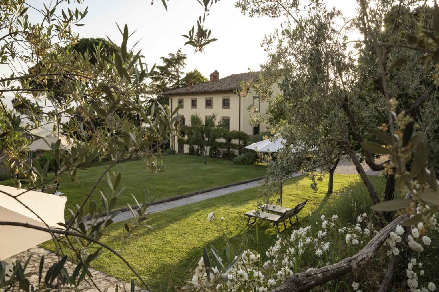 Scenic view of a countryside villa surrounded by lush greenery and olive trees, featuring a spacious garden with a sun loungers and an outdoor seating area. | Only on SopranoVillas