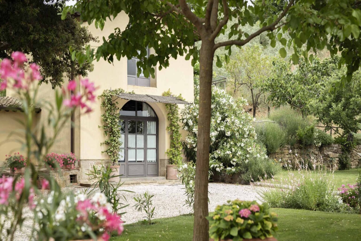 Charming countryside villa surrounded by lush greenery and vibrant flowers, featuring a welcoming entrance with large windows and a stone pathway. | Only on SopranoVillas