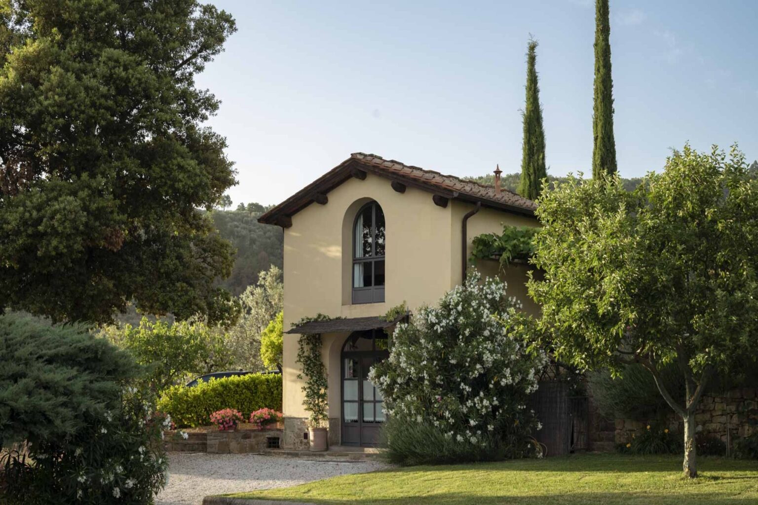 Charming countryside villa surrounded by lush greenery and tall cypress trees, featuring a welcoming entrance and blooming flowers in the garden. Ideal for nature lovers and serene getaways. | Only on SopranoVillas