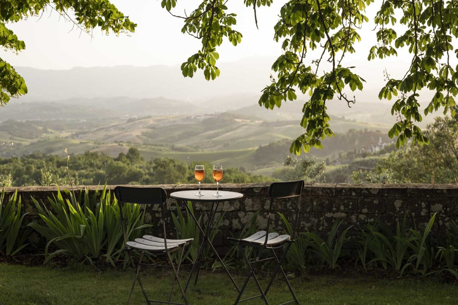 Scenic view of a tranquil outdoor setting featuring a small round table with two chairs, adorned with two glasses of drinks, overlooking lush green hills and valleys under a canopy of leaves. Perfect for relaxation and enjoying nature. | Only on SopranoVillas