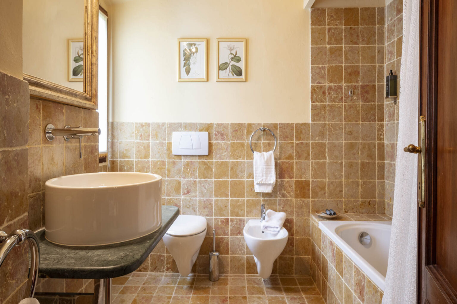 Modern bathroom interior featuring a round sink on a black countertop, a bidet, a toilet, and a bathtub, with decorative framed botanical prints on the wall and warm tile accents. | Only on SopranoVillas