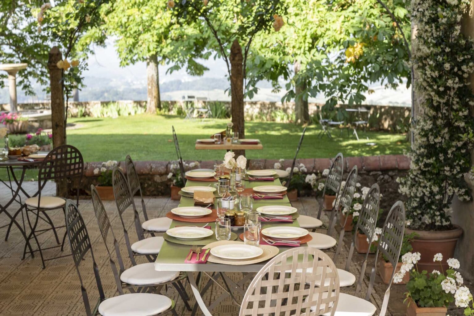 Outdoor dining setup with a long table elegantly arranged with plates and floral centerpieces, surrounded by lush greenery and trees, ideal for a garden party or event. | Only on SopranoVillas