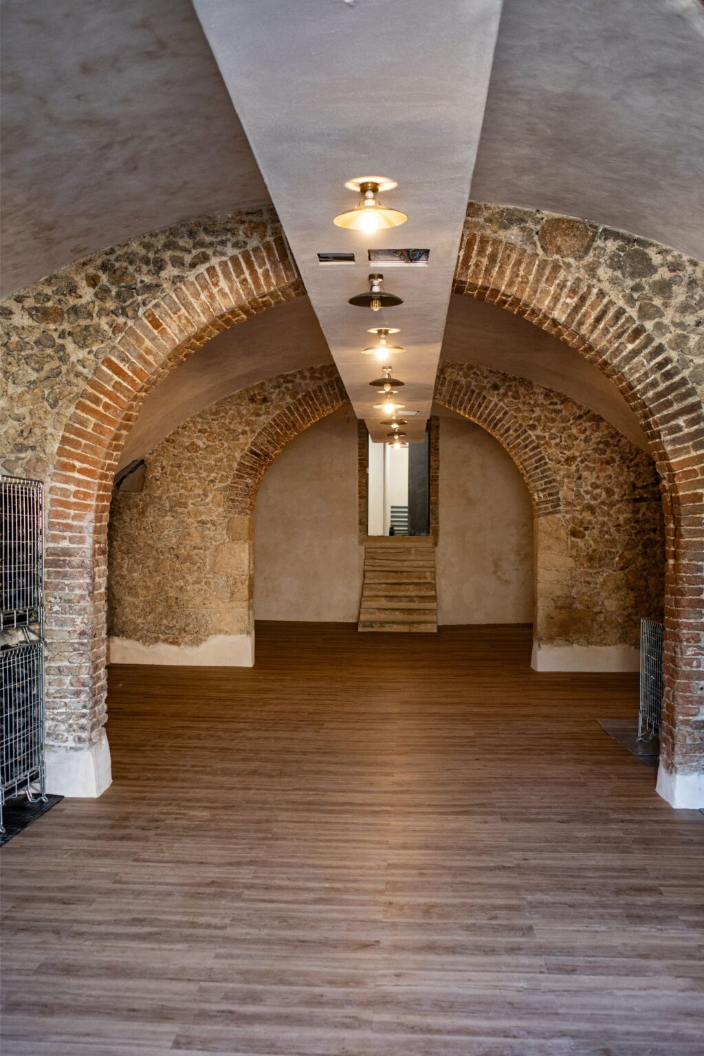 Interior view of a spacious, renovated room featuring arched brick walls and modern lighting. The wooden floor adds warmth, while a staircase at the end leads to another area. Metal grids are positioned along the sides, enhancing the industrial aesthetic. Ideal for events or as a creative workspace. | Only on SopranoVillas