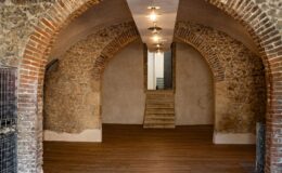 Interior view of a spacious, renovated room featuring arched brick walls and modern lighting. The wooden floor adds warmth, while a staircase at the end leads to another area. Metal grids are positioned along the sides, enhancing the industrial aesthetic. Ideal for events or as a creative workspace.