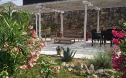 Outdoor garden seating area featuring wooden benches and a dining table under a pergola, surrounded by colorful flowers and lush greenery. Stone wall provides a rustic backdrop. Ideal for relaxation and gatherings.