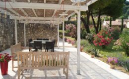 Outdoor patio with a wooden bench and dining table surrounded by vibrant flowers and greenery, featuring a stone wall and a pergola for shade. Ideal for relaxation and gatherings in a serene garden setting.