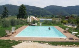 Scenic outdoor swimming pool surrounded by lush greenery and mountains, featuring sun loungers, umbrellas, and a couple enjoying the water on a sunny day. Ideal for relaxation and vacation getaways.