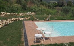 Serene outdoor pool area featuring a turquoise swimming pool, two white chairs, and a small table, surrounded by lush greenery and a stone border, ideal for relaxation and leisure.